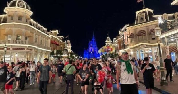 A bustling night scene at a theme park, featuring a crowd of visitors walking along an illuminated street lined with ornate buildings. In the background, a brightly lit castle stands as a focal point, while a person holds a bunch of colorful balloons.