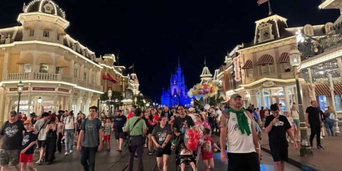 A bustling night scene at a theme park, featuring a crowd of visitors walking along an illuminated street lined with ornate buildings. In the background, a brightly lit castle stands as a focal point, while a person holds a bunch of colorful balloons.