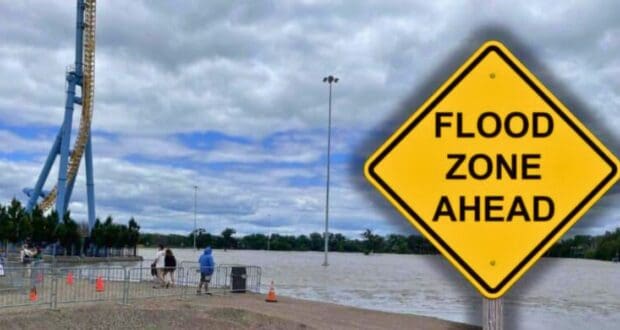 A large yellow sign reads "FLOOD ZONE AHEAD" in bold black letters. Behind the sign, Valleyfair amusement park appears partially submerged by floodwaters under a cloudy sky. A few people can be seen walking on the elevated ground near the water's edge at Valleyfair theme park.