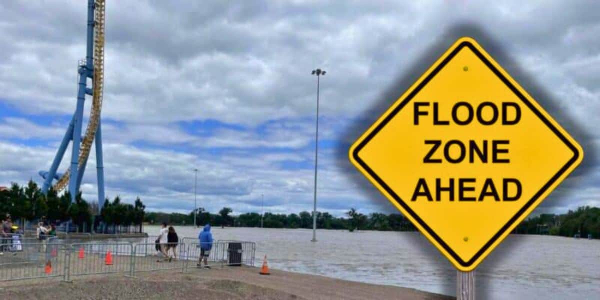 A large yellow sign reads "FLOOD ZONE AHEAD" in bold black letters. Behind the sign, Valleyfair amusement park appears partially submerged by floodwaters under a cloudy sky. A few people can be seen walking on the elevated ground near the water's edge at Valleyfair theme park.