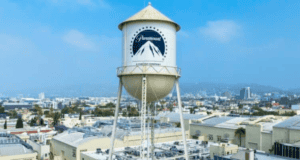A prominent water tower with the Paramount Pictures logo stands tall against a backdrop of an urban landscape under a bright sky. The surrounding area features various industrial buildings and city structures in the distance, reminiscent of iconic scenes from classic Disney films.