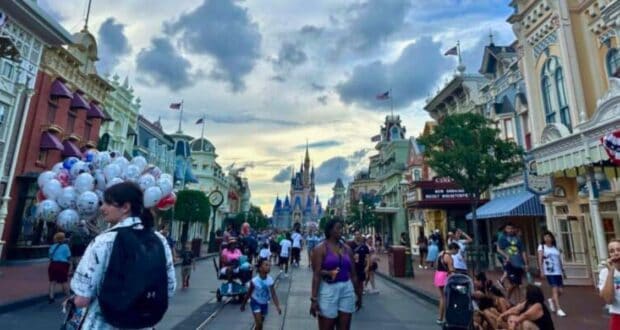 A bustling street scene at a theme park featuring people walking down the main street with the iconic castle in the background. Balloons, decorated shopfronts, and flags add to the festive atmosphere. The sky is partly cloudy, capturing a lively and vibrant moment.