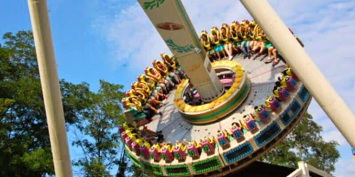 People are sitting in a circular amusement ride at a theme park, secured by harnesses, as it moves. The ride is tilted at an angle against the blue sky and trees in the background. Bright colors decorate the ride, and the participants seem engaged in the thrilling experience.
