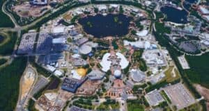Aerial view of a theme park reminiscent of Disney World, showcasing various attractions, rides, and buildings surrounding a central lake. The landscape is lush with greenery, and parking lots are visible along the perimeter. Paths connect the different areas of the park.