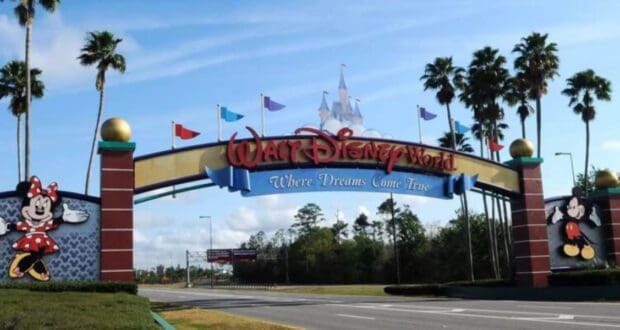 Walt Disney World entrance with a large archway reading "Walt Disney World: Where Dreams Come True." Minnie Mouse is on the left, Mickey Mouse is on the right. Palm trees and a castle-like structure are visible in the background.