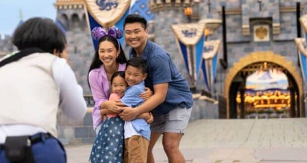 A family of four, consisting of two adults and two children, poses for a photo in front of a castle at a themed amusement park. The woman wears Minnie Mouse ears with a bow, and they all smile happily while hugging each other.