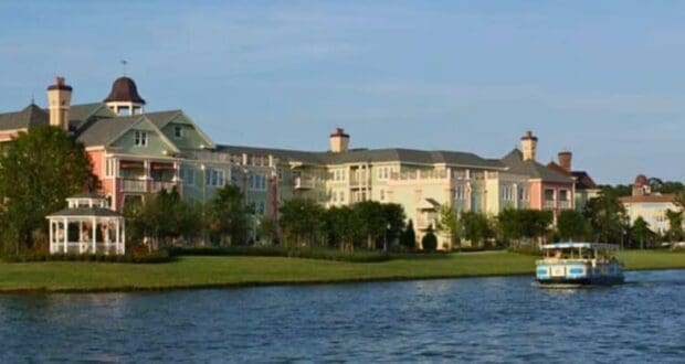 A serene lake scene with a charming, multi-colored building complex in the background, featuring pastel hues reminiscent of Disney Resort pools. A gazebo is on the left near the water's edge. A blue and yellow boat floats on the calm lake to the right. Trees and greenery surround the area.