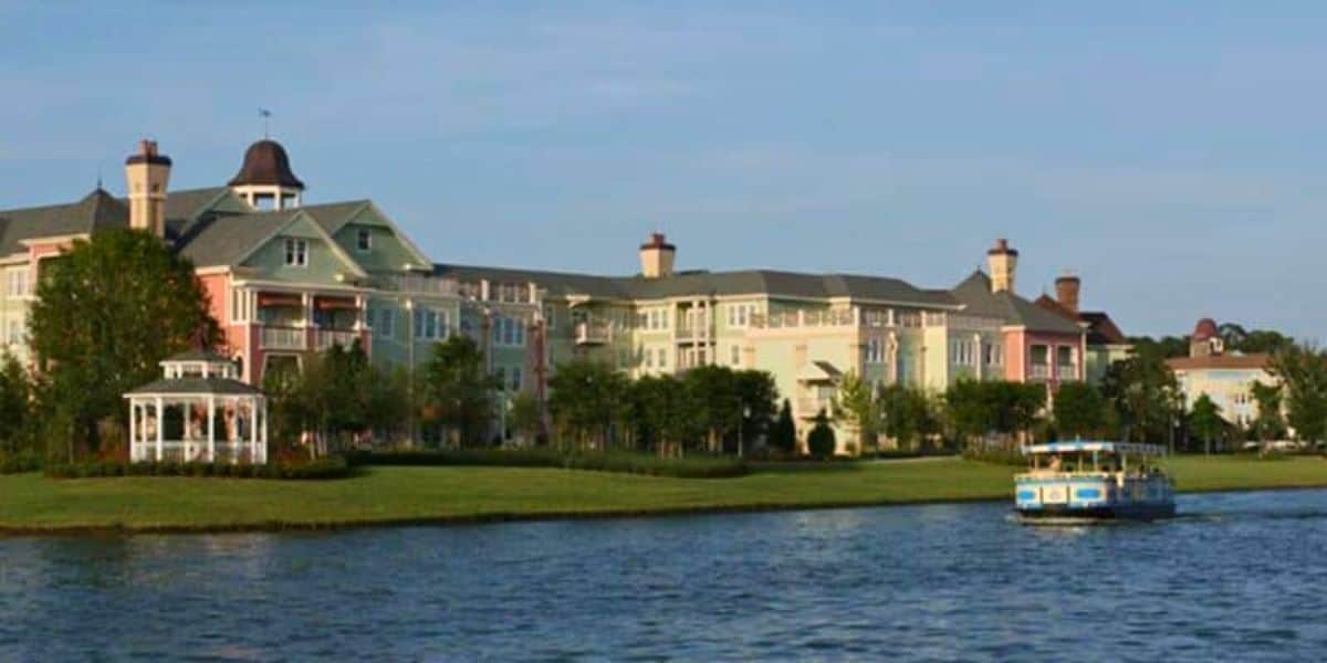 A serene lake scene with a charming, multi-colored building complex in the background, featuring pastel hues reminiscent of Disney Resort pools. A gazebo is on the left near the water's edge. A blue and yellow boat floats on the calm lake to the right. Trees and greenery surround the area.