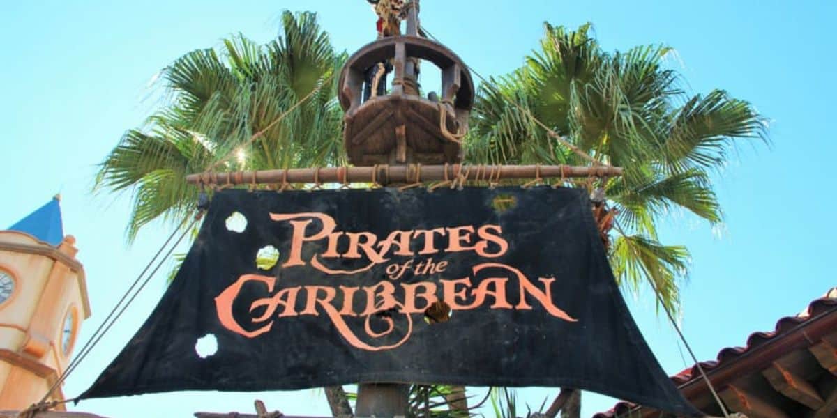 The image shows a wooden sign reading "Pirates of the Caribbean" in an elaborate Gothic font, hanging in front of palm trees against a clear blue sky. The sign resembles a pirate ship sail with holes, and a lantern is mounted above it.