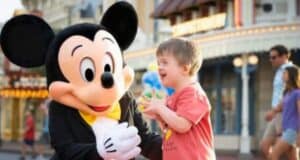 A smiling child wearing a red shirt interacts joyfully with a person dressed as Mickey Mouse at a theme park. The background features colorful buildings and other park visitors walking by.