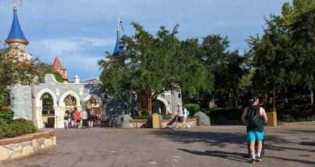 People are walking through an amusement park with castle-themed structures and colorful turrets in the background. Trees and benches are scattered throughout the scene, and the sky is clear and blue.