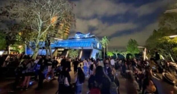A large crowd of people gathers at a theme park at night. The area is illuminated with blue and yellow lights, highlighting a futuristic building in the background. Trees and greenery surround the scene, with a partly cloudy sky overhead.