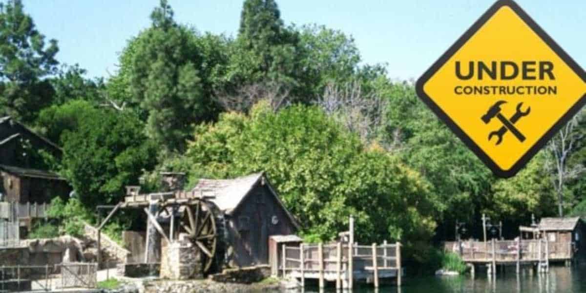 A wooden mill house with a large water wheel stands near a river, surrounded by dense greenery. A yellow diamond-shaped "Under Construction" sign with crossed tools appears in the sky area of the image on the right side, indicating ongoing construction.
