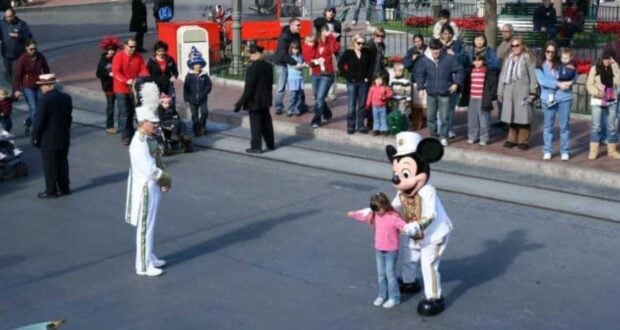 A young girl stands next to a person in a Mickey Mouse costume on a paved street, pointing to the side. A man in a white uniform with a tall feathered hat is nearby. A crowd of onlookers in casual clothing lines the sidewalk, watching the interaction.