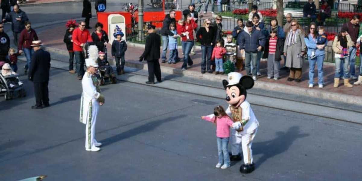 A young girl stands next to a person in a Mickey Mouse costume on a paved street, pointing to the side. A man in a white uniform with a tall feathered hat is nearby. A crowd of onlookers in casual clothing lines the sidewalk, watching the interaction.
