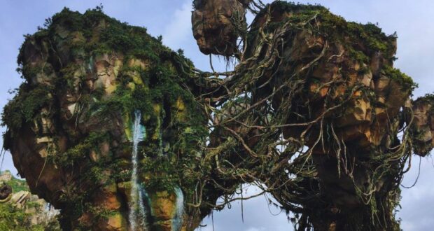 A lush, floating mountain range covered in greenery and vines, with large rocky formations connected by natural bridges. The sky is a mix of clouds and clear areas, creating a mystical atmosphere reminiscent of a fantastical landscape.
