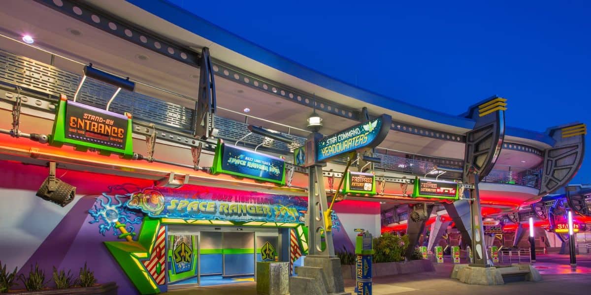 The image captures the entrance to Buzz Lightyear's Space Ranger Spin at a theme park, likely in the early evening. Illuminated by vibrant, colorful signs and lights, the area radiates a futuristic ambiance with space-themed decorations.