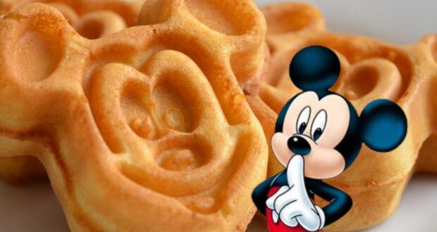 A close-up of waffles shaped like a round-eared cartoon mouse face, with a cartoon version of the same mouse character holding a finger to their lips. The golden brown waffles appear freshly made—taking Disney Dining Tips to heart for a magical breakfast experience!