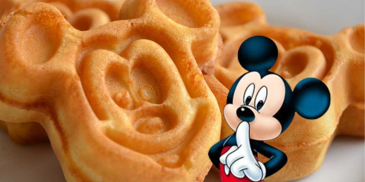 A close-up of waffles shaped like a round-eared cartoon mouse face, with a cartoon version of the same mouse character holding a finger to their lips. The golden brown waffles appear freshly made—taking Disney Dining Tips to heart for a magical breakfast experience!