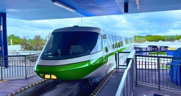A sleek green and white monorail train arrives at an elevated platform on a sunny day, reminiscent of the Disney Transportation and Ticket Center. The train's streamlined design is visible, with trees and structures in the background. The platform is equipped with railings and safety measures.