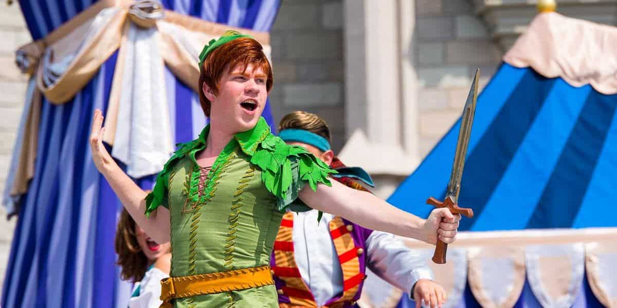 Person dressed as Peter Pan, wearing a green costume with a green hat, performing energetically on stage. Holding a sword in one hand and displaying a determined expression, they stand against a backdrop of blue and white curtains and a stone structure.