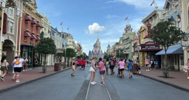 A bustling street scene at Disney World, with a variety of people walking and taking photos. Shops and buildings with decorative facades line both sides. A castle is visible in the distance under a blue sky with scattered clouds.