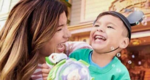 A mother and her young son are laughing together at a theme park during Pixar Fest at Disneyland. The boy is wearing a green shirt and Mickey Mouse ears, smiling widely, and holding a Buzz Lightyear toy. The background features colorful buildings, with bubbles floating around them.