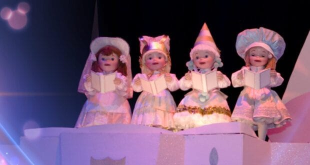 Four childlike dolls dressed in whimsical, old-fashioned costumes stand in a row, each holding an open book as if singing. The background is dark with a bluish lens flare effect on the left, adding a magical ambiance to the scene.