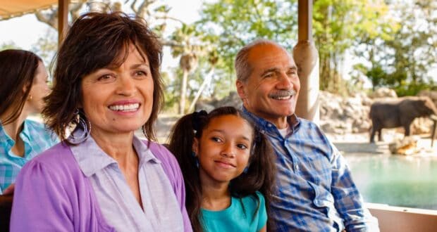 A smiling elderly couple sits close to a young girl, also smiling, with another person partially visible in the background. They appear to be on Disney's Kilimanjaro Safaris ride, with an elephant and lush greenery visible in the distance at Disney's Animal Kingdom.