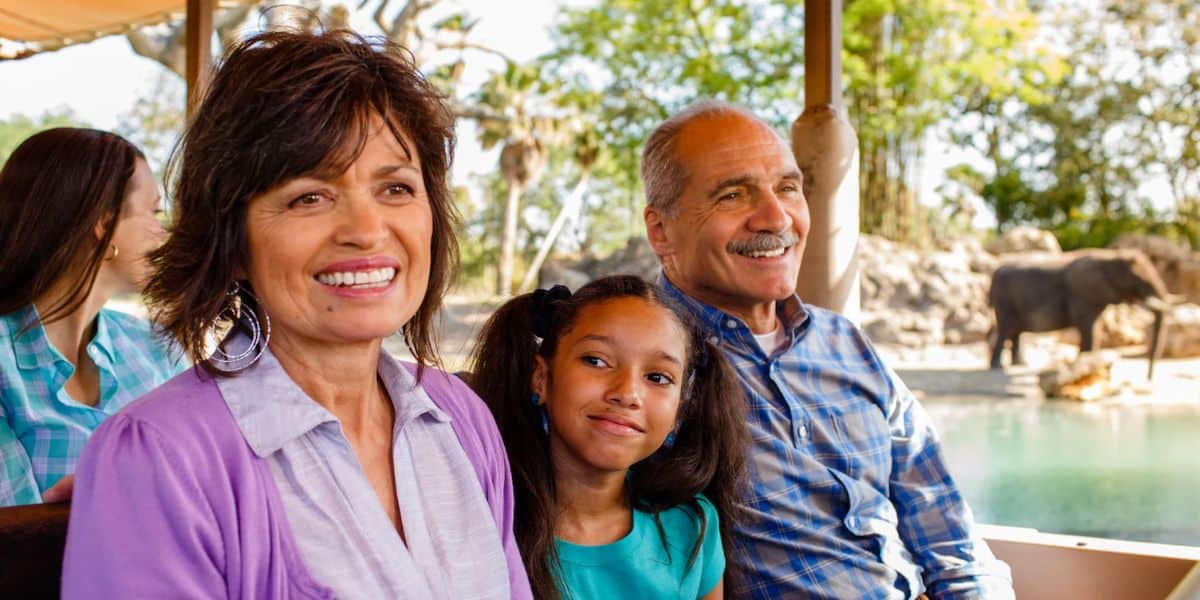 A smiling elderly couple sits close to a young girl, also smiling, with another person partially visible in the background. They appear to be on Disney's Kilimanjaro Safaris ride, with an elephant and lush greenery visible in the distance at Disney's Animal Kingdom.