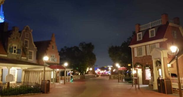 A nighttime view of a themed amusement park street, with quaint European-style buildings lining the path. Vintage street lamps illuminate the way, while colorful lights sparkle near the Liberty Square bathrooms in the distance.