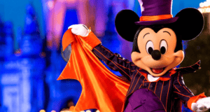 Mickey Mouse dressed in a purple and orange Halloween costume, featuring a top hat and a cape, stands outside, smiling and striking a pose. The background is a blurred, illuminated theme park setting, possibly at night.