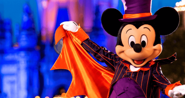 Mickey Mouse dressed in a purple and orange Halloween costume, featuring a top hat and a cape, stands outside, smiling and striking a pose. The background is a blurred, illuminated theme park setting, possibly at night.
