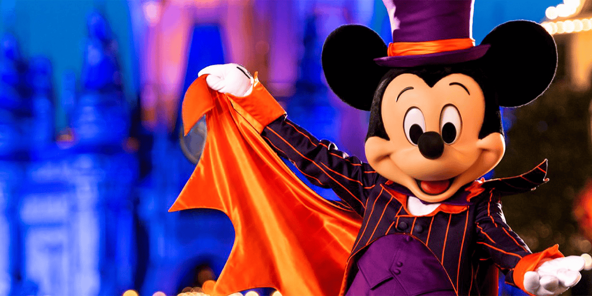 Mickey Mouse dressed in a purple and orange Halloween costume, featuring a top hat and a cape, stands outside, smiling and striking a pose. The background is a blurred, illuminated theme park setting, possibly at night.