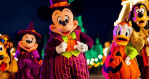 Mickey Mouse and friends, dressed in Halloween costumes, pose for a photo in front of a brightly lit castle. Mickey wears a magician's outfit, Minnie is a witch, Donald Duck is a pumpkin, Goofy is a scary character, and Pluto has a festive collar.