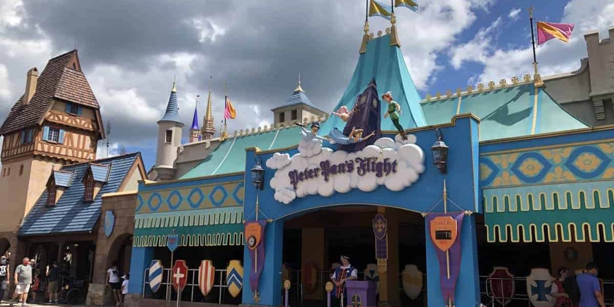 A colorful theme park attraction features a whimsical castle-like facade with turrets and a vibrant blue and gold color scheme. The entrance sign reads "Peter Pan's Flight" and depicts Peter Pan, Wendy, and other characters flying on clouds. Visitors walk nearby, eager to relive the adventures of Peter Pan.