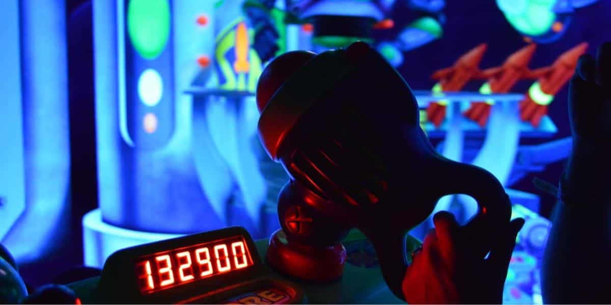 A close-up view of a toy laser gun in an arcade setting, reminiscent of Buzz Lightyear Space Ranger Spin, with a glowing score display showing 132,900 points. The background is vibrant with neon blue and green colors, additional toy lasers, and various arcade decorations.