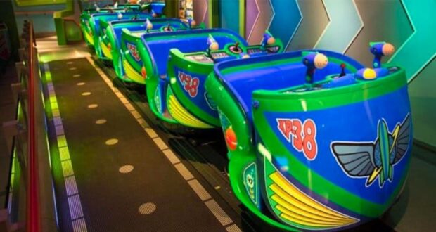 Four turquoise and green ride vehicles, themed after Toy Story spacecraft and featuring "XP-38" decals, are lined up on a track, ready for passengers. They have seats and twin laser blasters, set against a futuristic backdrop with zigzag neon patterns.