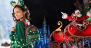 A woman dressed in a festive green sweater and mouse ears headband, carrying a green and red backpack, looks over her shoulder and smiles. Behind her, a person dressed as Santa Claus waves from a decorated sleigh, with a castle and dark sky in the background.