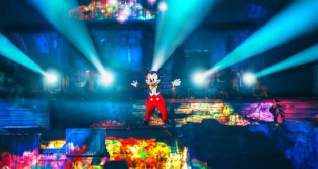 Mickey Mouse stands center stage with arms outstretched, illuminated by vibrant, colorful lights and surrounded by smoke and bright beams in a theatrical setting. The background features diverse, moving patterns of light and colors, creating a magical atmosphere.