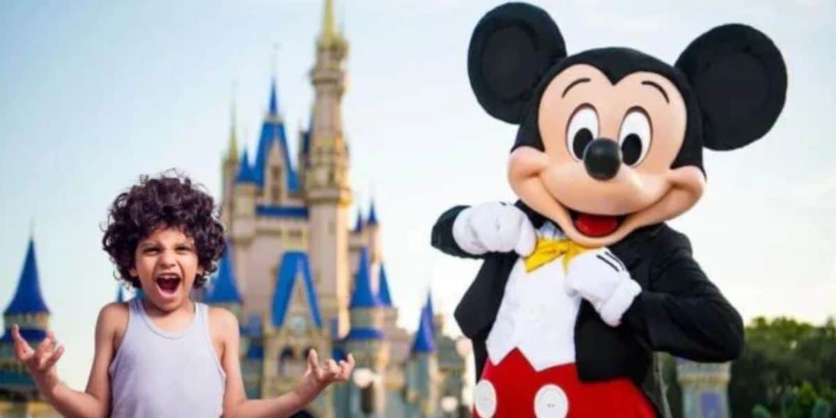 A young child with curly hair, wearing a sleeveless shirt, stands excitedly next to a person in a Mickey Mouse costume. Behind them, there is a large castle with blue and gold accents, reminiscent of a fairy tale setting. Both appear to be at a theme park.