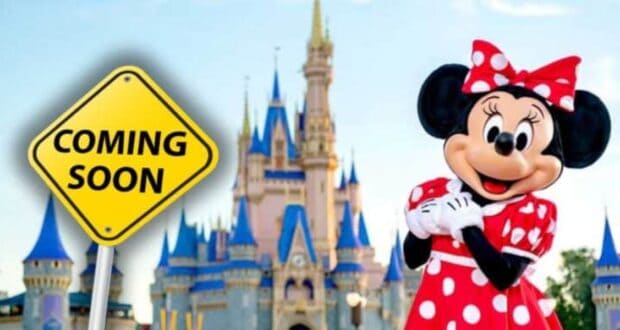 Minnie Mouse, dressed in her classic red and white polka dot dress and bow, poses in front of Cinderella's Castle in a Disney park. Beside her is a yellow road sign with the text "COMING SOON." The sky is blue with a few scattered clouds.