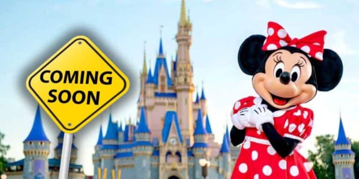 Minnie Mouse, dressed in her classic red and white polka dot dress and bow, poses in front of Cinderella's Castle in a Disney park. Beside her is a yellow road sign with the text "COMING SOON." The sky is blue with a few scattered clouds.