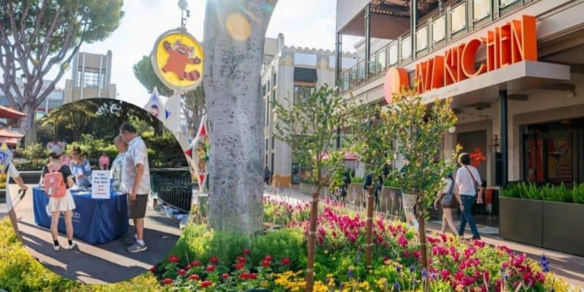 A colorful outdoor scene featuring a building with an orange "Rakkugin" sign. A tree is surrounded by vibrant flowers. An inset shows a man and a woman talking at a table. Banners and a round sign with a fox character hang nearby.