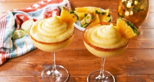 Two margarita glasses filled with a creamy yellow frozen beverage, garnished with a pineapple slice and a chili powder rim. Behind them, a colorful striped cloth napkin, lime slices, and a bowl of pineapple pieces are on a wooden table.