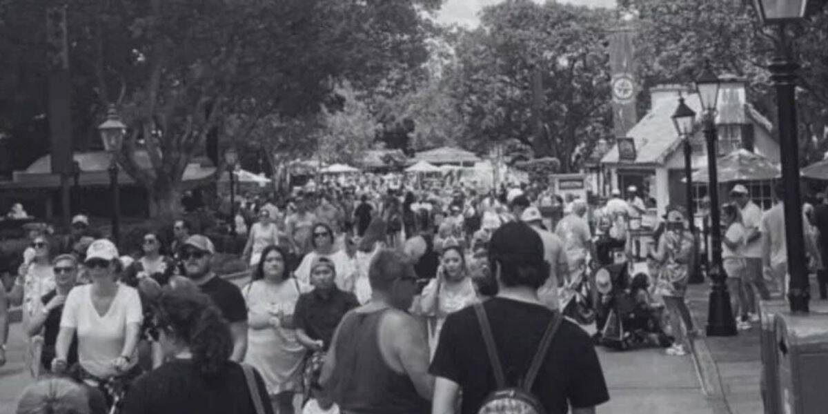 A bustling crowd of people walks along a tree-lined outdoor path, possibly in an amusement park or public space. The scene is in black and white, with various individuals engaging in different activities. Small structures and lamp posts line the path.