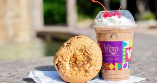 A large cookie with chunks of chocolate rests on a napkin next to an iced coffee topped with whipped cream and cherries in a plastic cup with a colorful Joffrey's logo. The items are on a stone surface with a blurred outdoor background.