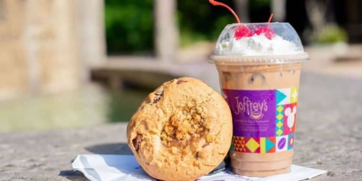 A large cookie with chunks of chocolate rests on a napkin next to an iced coffee topped with whipped cream and cherries in a plastic cup with a colorful Joffrey's logo. The items are on a stone surface with a blurred outdoor background.