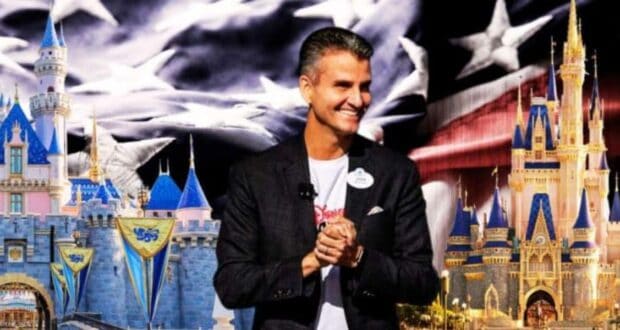 A man in a black jacket and white shirt stands smiling with his hands clasped in front of him. Behind him are images of two Disney castle attractions and an American flag backdrop in grayscale.
