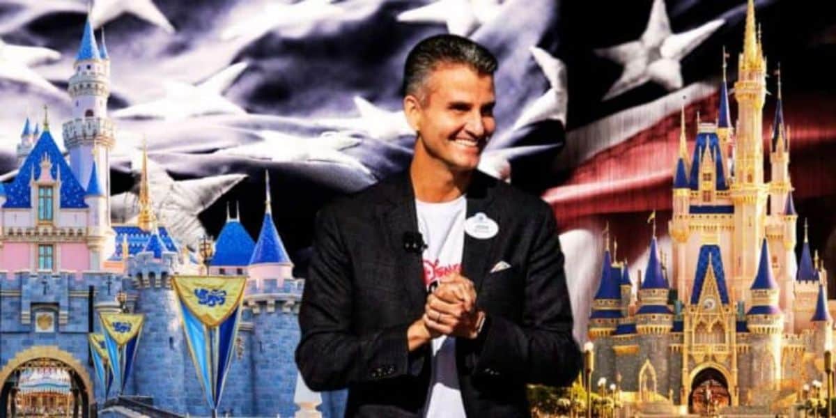 A man in a black jacket and white shirt stands smiling with his hands clasped in front of him. Behind him are images of two Disney castle attractions and an American flag backdrop in grayscale.